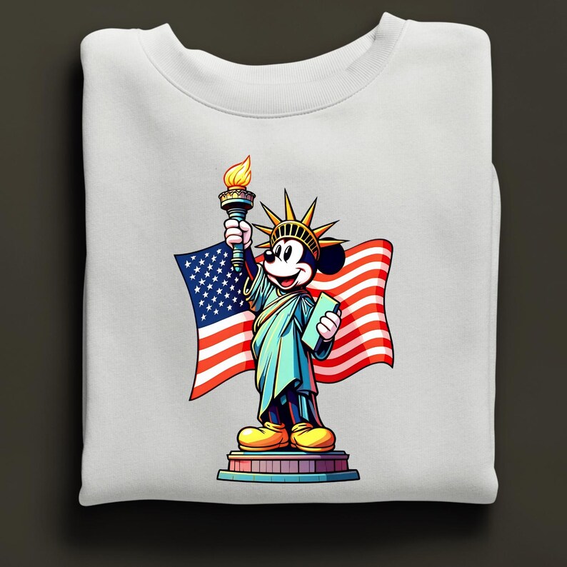 Puede incluir: Sudadera blanca con una representaci&oacute;n de dibujos animados de Mickey Mouse como la Estatua de la Libertad, sosteniendo una antorcha y una tableta, con una bandera estadounidense de fondo. El dise&ntilde;o es colorido y patri&oacute;tico.
