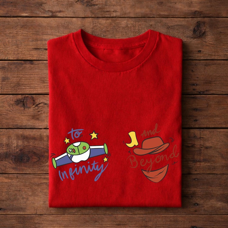 Op de afbeelding: Een rood t-shirt met een cartoonontwerp en de tekst "to infinity and beyond". Het ontwerp bevat een ruimteschip, een cowboyhoed en een glimlach. Het shirt is op een houten oppervlak gevouwen.