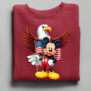 May include: A maroon sweatshirt featuring a graphic of Mickey Mouse holding an American flag, with a bald eagle behind him. The eagle has brown wings and a white head. Mickey is wearing red shorts, yellow shoes, and white gloves.