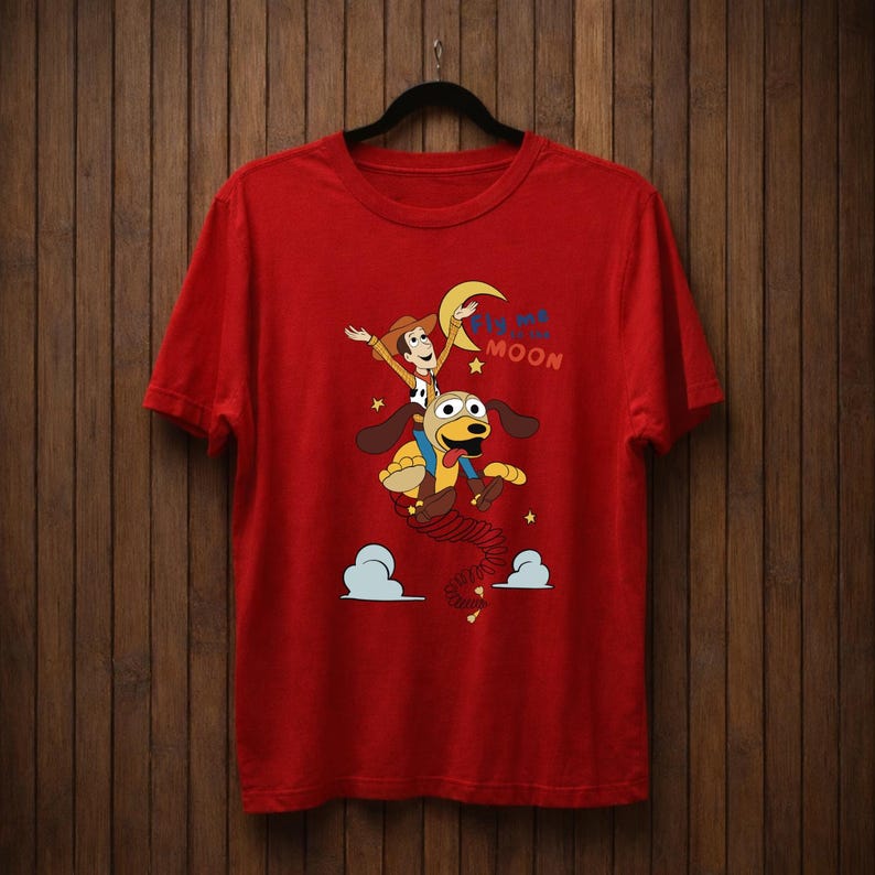 Peut inclure: T-shirt rouge avec un dessin anim&eacute; d'un cow-boy chevauchant un chien vers la lune. Le graphisme comprend le texte "To the Moon". Le t-shirt est suspendu &agrave; un cintre noir.