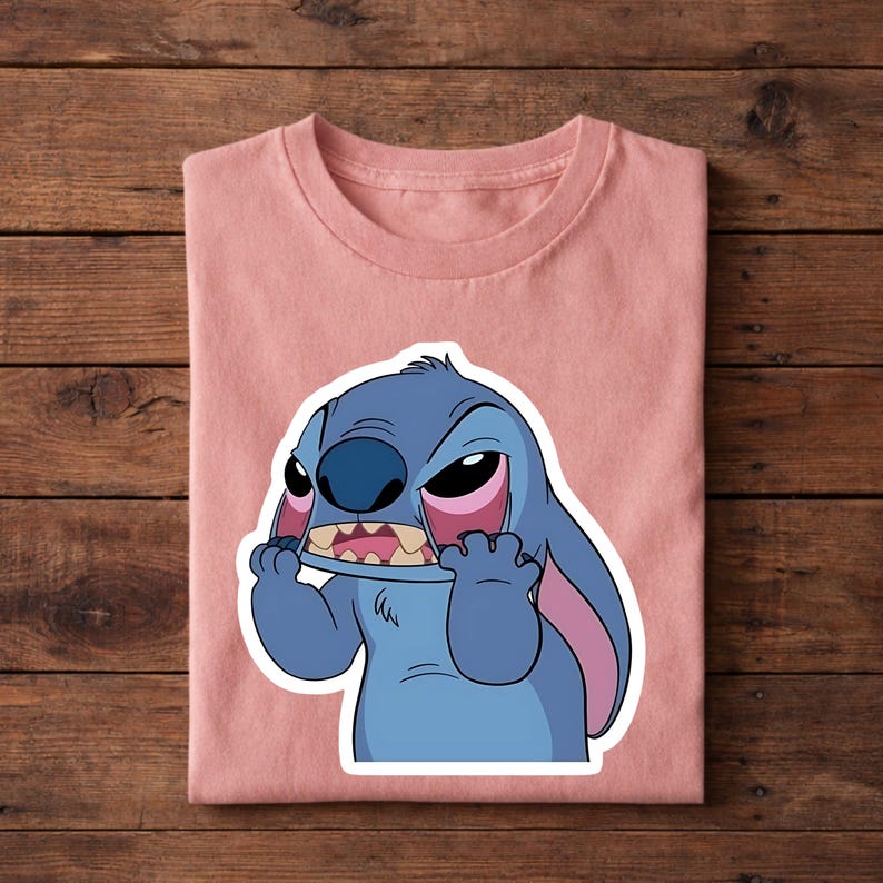 May include: A folded, light pink t-shirt featuring a cartoon image of Stitch from Lilo & Stitch. The blue alien character has an angry expression, with pink accents on his ears and eyes. The shirt is made of soft material.