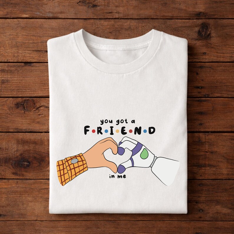 May include: White t-shirt with a graphic design featuring the text "you got a FRIEND in me" in colorful letters. The design includes cartoon hands forming a heart shape, one with a plaid sleeve and the other with a purple and green design.