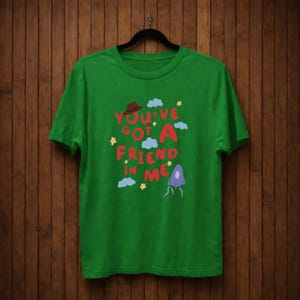 Op de afbeelding: Een groen t-shirt met de tekst "You've Got A Friend In Me" in rood, roze en blauw. Het ontwerp bevat een bruine hoed, wolken, sterren en een paars, buitenaards figuur. Het t-shirt hangt aan een zwarte hanger.