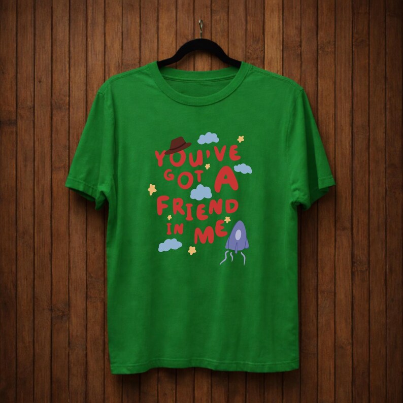 Op de afbeelding: Een groen t-shirt met de tekst "You've Got A Friend In Me" in rood, roze en blauw. Het ontwerp bevat een bruine hoed, wolken, sterren en een paars, buitenaards figuur. Het t-shirt hangt aan een zwarte hanger.