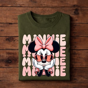 K&ouml;nnte beinhalten: Olivgr&uuml;nes T-Shirt mit einer Minnie Mouse-Grafik, die eine herzf&ouml;rmige Sonnenbrille und eine rosa Schleife tr&auml;gt. Das Wort "MINNIE" ist vertikal in Rosa gedruckt. Das T-Shirt ist auf einer Holzoberfl&auml;che gefaltet.