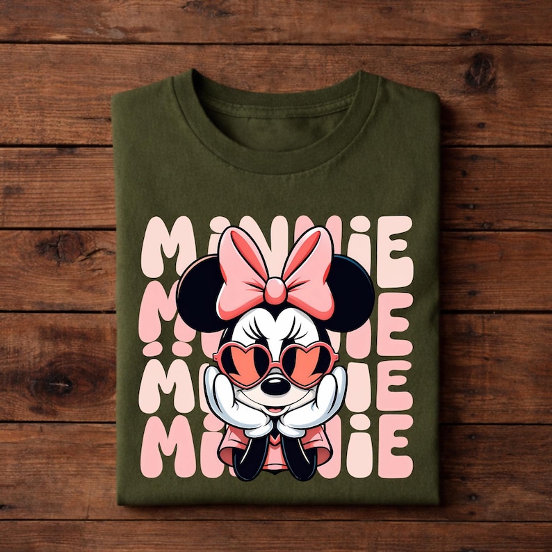 K&ouml;nnte beinhalten: Olivgr&uuml;nes T-Shirt mit einer Minnie Mouse-Grafik, die eine herzf&ouml;rmige Sonnenbrille und eine rosa Schleife tr&auml;gt. Das Wort "MINNIE" ist vertikal in Rosa gedruckt. Das T-Shirt ist auf einer Holzoberfl&auml;che gefaltet.