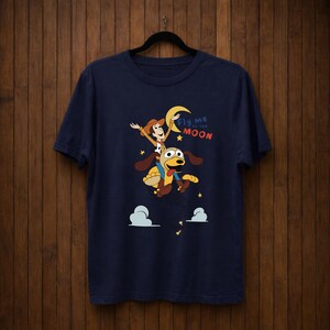 Peut inclure: T-shirt bleu marine avec une illustration de dessin anim&eacute; d'un personnage de cow-boy chevauchant un chien vers la lune. Le texte "Fly me to the Moon" est &eacute;galement pr&eacute;sent. Le design comprend des &eacute;toiles et des nuages.