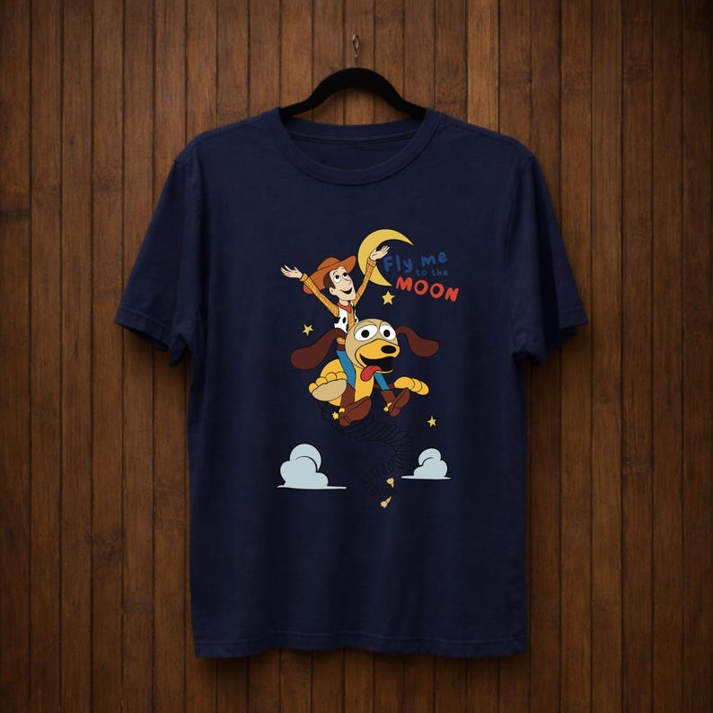 Peut inclure: T-shirt bleu marine avec une illustration de dessin anim&eacute; d'un personnage de cow-boy chevauchant un chien vers la lune. Le texte "Fly me to the Moon" est &eacute;galement pr&eacute;sent. Le design comprend des &eacute;toiles et des nuages.