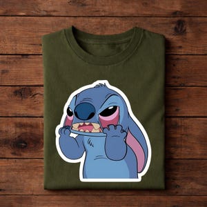May include: Olive green t-shirt featuring a cartoon illustration of Stitch from Lilo & Stitch. The character is blue with large ears, a wide mouth, and pink accents. The design is centered on the shirt.