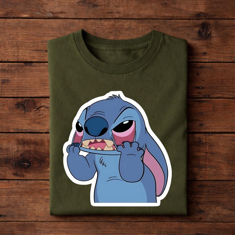 May include: Olive green t-shirt featuring a cartoon illustration of Stitch from Lilo & Stitch. The character is blue with large ears, a wide mouth, and pink accents. The design is centered on the shirt.