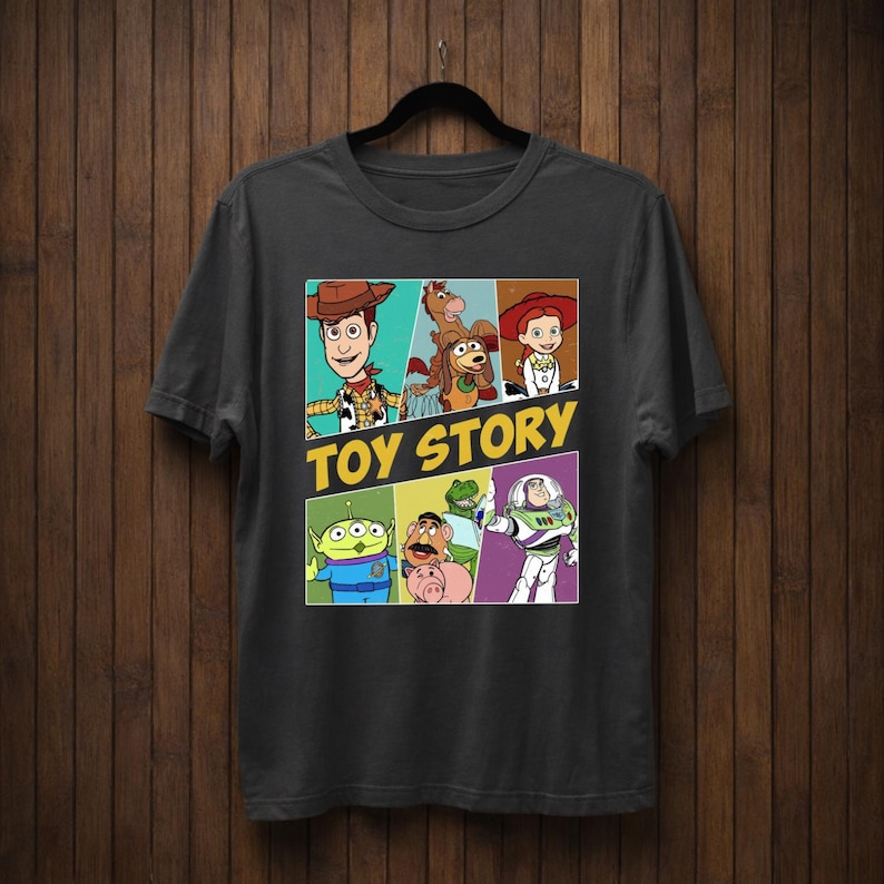 K&ouml;nnte beinhalten: Dunkelgraues T-Shirt mit einer farbenfrohen Grafik von Charakteren aus dem Animationsfilm "Toy Story". Das Design zeigt Woody, Buzz Lightyear und andere Charaktere mit den Worten "TOY STORY" in Gelb.