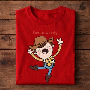 Puede incluir: Camiseta roja con una ilustraci&oacute;n de dibujos animados de un personaje de vaquero con sombrero marr&oacute;n, camisa amarilla, pantalones azules y botas marrones. El texto "Feelin woody" est&aacute; encima del personaje. El personaje hace un signo de paz.