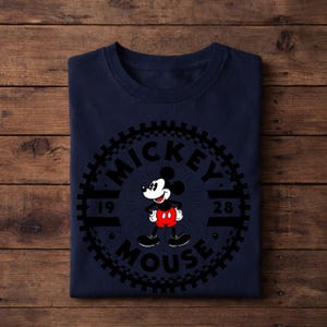 K&ouml;nnte beinhalten: Marineblaues T-Shirt mit einem kreisf&ouml;rmigen Mickey Mouse-Motiv. Die Grafik enth&auml;lt die Worte "Mickey Mouse" und das Jahr "1928". Mickey Mouse wird in seiner klassischen Pose mit roter Hose und wei&szlig;en Handschuhen dargestellt.