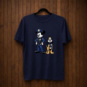 May include: Navy blue t-shirt featuring a cartoon illustration of Mickey Mouse and Pluto dressed as police officers. Mickey Mouse is wearing a police uniform and hat, while Pluto has a police badge. The design is centered on the front of the shirt.
