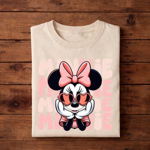 K&ouml;nnte beinhalten: Ein gefaltetes, cremefarbenes T-Shirt mit einem Minnie Mouse-Motiv. Minnie ist mit einer rosa Schleife, herzf&ouml;rmigen Sonnenbrillen und einem rosa Kleid dargestellt. Das Wort "MINNIE" wird vertikal in Rosa wiederholt.