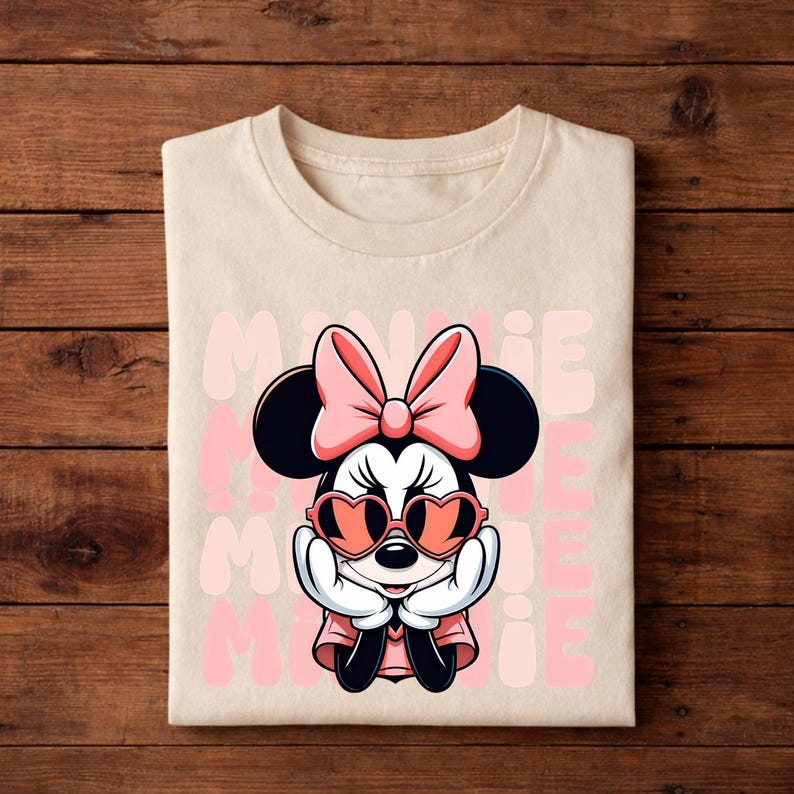 K&ouml;nnte beinhalten: Ein gefaltetes, cremefarbenes T-Shirt mit einem Minnie Mouse-Motiv. Minnie ist mit einer rosa Schleife, herzf&ouml;rmigen Sonnenbrillen und einem rosa Kleid dargestellt. Das Wort "MINNIE" wird vertikal in Rosa wiederholt.