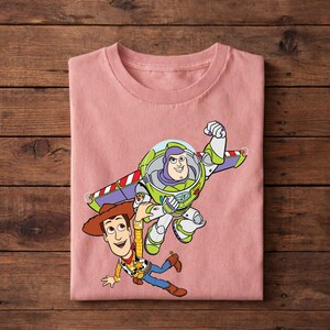 May include: A light pink t-shirt featuring a graphic of Buzz Lightyear and Woody from Toy Story. Buzz Lightyear is in his space suit with wings extended, and Woody is in his cowboy outfit. The design is colorful and cartoon-like.