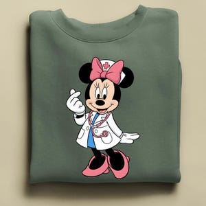 Puede incluir: Sudadera verde con Minnie Mouse de dibujos animados vestida de m&eacute;dico. Minnie Mouse lleva una bata blanca, un lazo rosa y un estetoscopio. Est&aacute; haciendo un coraz&oacute;n con los dedos. La sudadera est&aacute; doblada.