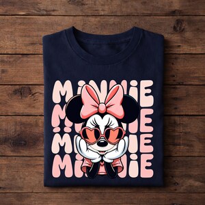 K&ouml;nnte beinhalten: Marineblaues T-Shirt mit einem Minnie Mouse-Motiv. Das Design zeigt Minnie mit herzf&ouml;rmiger Sonnenbrille, einer rosa Schleife und dem Namen "Minnie" vertikal in Rosa wiederholt. Das T-Shirt ist auf einer Holzoberfl&auml;che gefaltet.