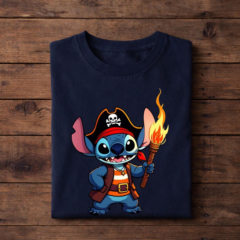 Pode incluir: Camiseta azul marinho com um personagem de desenho animado vestido de pirata. O personagem azul tem olhos grandes, um chap&eacute;u de pirata com uma caveira e ossos cruzados e uma tocha com uma chama. A camiseta est&aacute; dobrada em uma superf&iacute;cie de madeira.