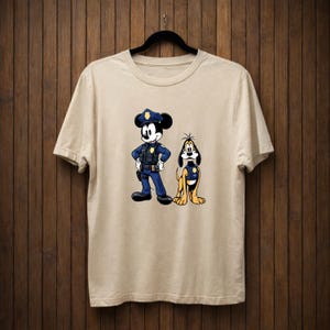 May include: Beige t-shirt featuring a cartoon illustration of Mickey Mouse and Pluto dressed as police officers. Mickey Mouse is in a blue uniform with a police hat, and Pluto is in a yellow uniform.