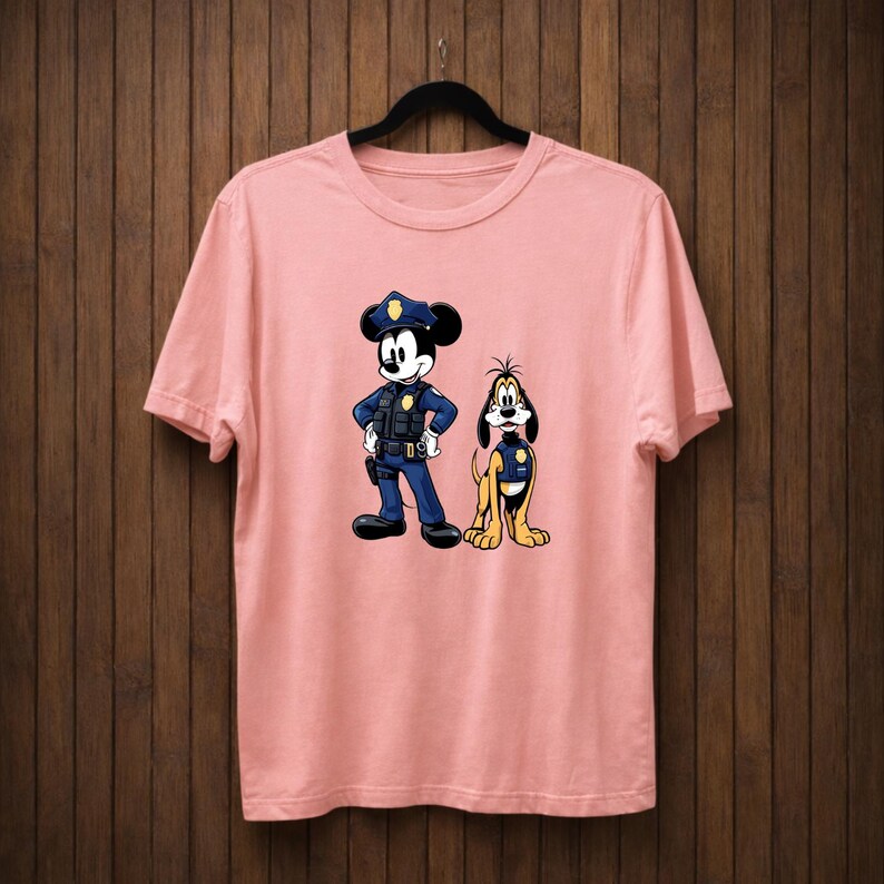 May include: A light pink t-shirt featuring a cartoon illustration of Mickey Mouse and Pluto dressed as police officers. Mickey Mouse is wearing a blue police uniform and hat. Pluto is wearing a yellow police vest. The shirt is hanging on a black hanger.