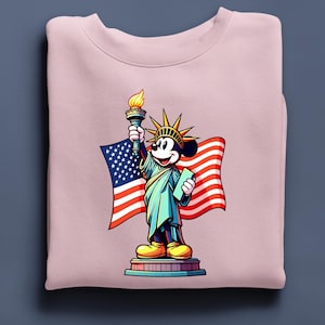 Puede incluir: Sudadera rosa claro con una ilustraci&oacute;n de dibujos animados de Mickey Mouse como la Estatua de la Libertad, sosteniendo una antorcha y una tableta, con una bandera estadounidense de fondo. El dise&ntilde;o es colorido y patri&oacute;tico.