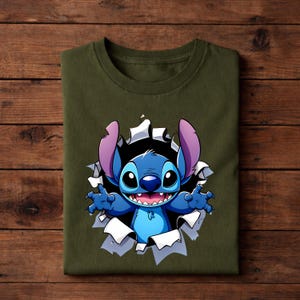 May include: Olive green t-shirt featuring a graphic of the character Stitch bursting through a torn hole. Stitch is blue with large ears, a wide smile, and outstretched arms. The design is centered on the folded shirt.