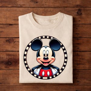 May include: A folded, cream-colored t-shirt featuring a smiling Mickey Mouse graphic. The cartoon character is framed by a black and white striped circle. Mickey Mouse is wearing a red and black outfit with white gloves.