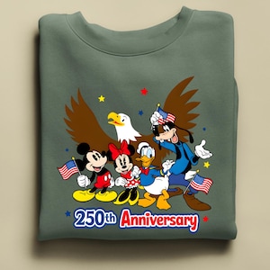 Puede incluir: Una sudadera verde salvia con Mickey Mouse, Minnie Mouse, Donald Duck y Goofy con banderas americanas y un &aacute;guila calva. El texto "250th Anniversary" est&aacute; en rojo y azul.