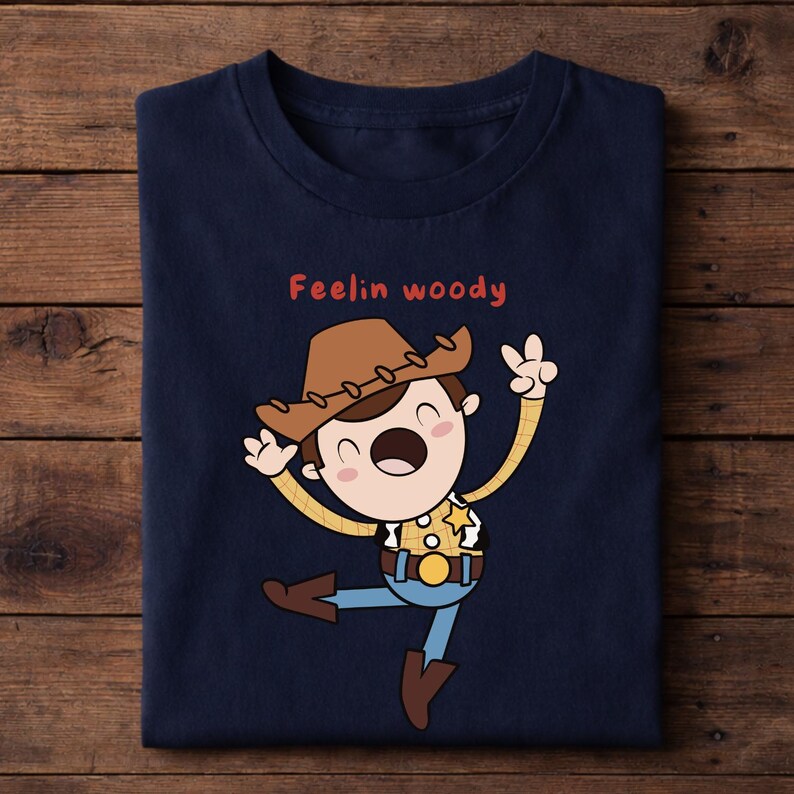 Puede incluir: Camiseta azul marino con un personaje de dibujos animados de vaquero con sombrero marr&oacute;n, camisa amarilla, vaqueros azules y una placa de sheriff. El texto rojo "Feelin woody" est&aacute; encima del personaje. La camiseta est&aacute; doblada sobre una superficie de madera.