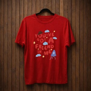Op de afbeelding: Rood t-shirt met de tekst "You've Got A Friend In Me" in een speels lettertype. Het ontwerp bevat een bruine hoed, blauwe wolken, gele sterren en een blauwe raket. Het shirt hangt aan een zwarte hanger.