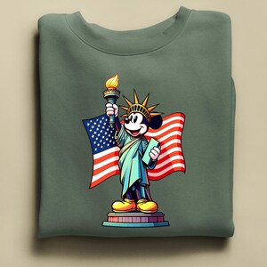 Puede incluir: Una sudadera verde salvia con una representaci&oacute;n de dibujos animados de Mickey Mouse como la Estatua de la Libertad. Mickey sostiene una antorcha y una tableta, con una bandera estadounidense de fondo. El dise&ntilde;o es colorido y patri&oacute;tico.
