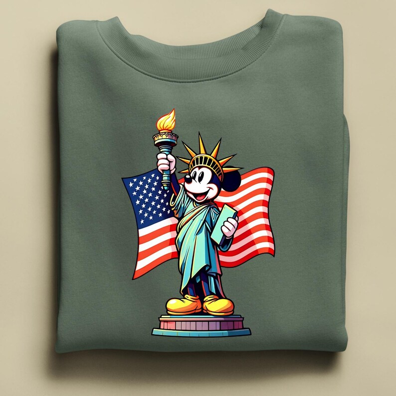 Puede incluir: Una sudadera verde salvia con una representaci&oacute;n de dibujos animados de Mickey Mouse como la Estatua de la Libertad. Mickey sostiene una antorcha y una tableta, con una bandera estadounidense de fondo. El dise&ntilde;o es colorido y patri&oacute;tico.