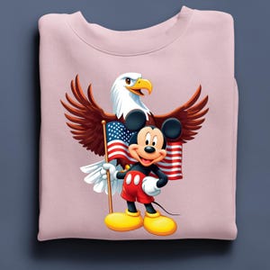 May include: A light pink sweatshirt featuring a graphic of Mickey Mouse holding an American flag, with a bald eagle behind him. Mickey is wearing red shorts, black gloves, and yellow shoes. The eagle has brown wings and a white head.