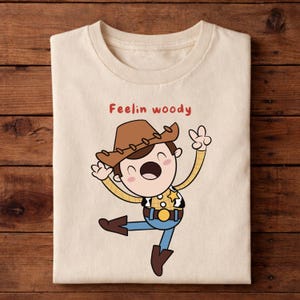 Puede incluir: Camiseta crema doblada con una ilustraci&oacute;n de dibujos animados de un personaje de vaquero con sombrero marr&oacute;n, camisa amarilla, pantalones azules y botas marrones. El texto "Feelin woody" est&aacute; impreso en rojo.