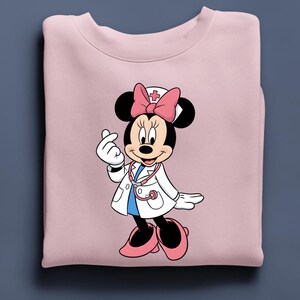 Puede incluir: Sudadera rosa claro con un dibujo de Minnie Mouse vestida de enfermera. Minnie Mouse lleva una bata blanca, un lazo rosa y un estetoscopio. El dibujo est&aacute; centrado en la sudadera.