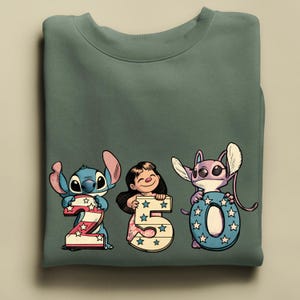 Puede incluir: Sudadera verde salvia con los personajes Stitch, Lilo y Angel sosteniendo los n&uacute;meros 2, 5 y 0. Los n&uacute;meros est&aacute;n decorados con estrellas y patrones patri&oacute;ticos. La sudadera est&aacute; doblada.