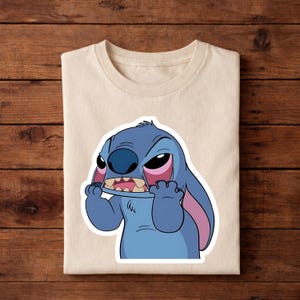 May include: A cream-colored t-shirt featuring a cartoon image of Stitch from Lilo & Stitch. The blue alien character has an angry expression, with pink accents on his ears and eyes. The shirt is folded on a wooden surface.
