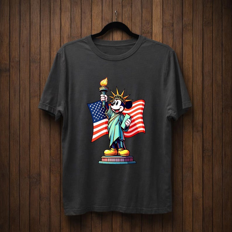 May include: Dark gray t-shirt featuring a cartoon character dressed as the Statue of Liberty, holding a torch and an American flag. The character has a yellow crown and shoes. The design is centered on the shirt.