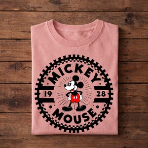 K&ouml;nnte beinhalten: Ein gefaltetes, hellrosa T-Shirt mit einer schwarz-wei&szlig;en Grafik von Mickey Mouse. Die Grafik zeigt die Worte "Mickey Mouse" und die Zahlen "19" und "28" in einem kreisf&ouml;rmigen Design. Das T-Shirt ist aus weichem Material.