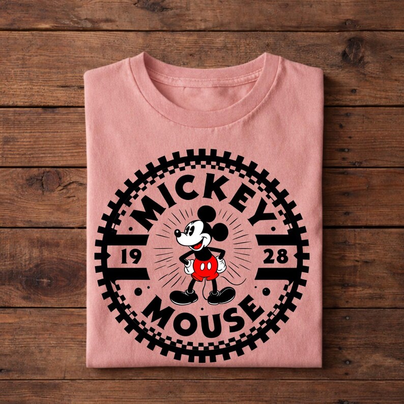 K&ouml;nnte beinhalten: Ein gefaltetes, hellrosa T-Shirt mit einer schwarz-wei&szlig;en Grafik von Mickey Mouse. Die Grafik zeigt die Worte "Mickey Mouse" und die Zahlen "19" und "28" in einem kreisf&ouml;rmigen Design. Das T-Shirt ist aus weichem Material.