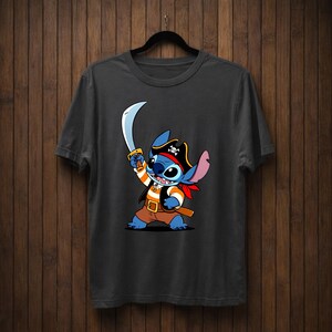 May include: Dark gray t-shirt featuring a cartoon Stitch character dressed as a pirate. Stitch is blue with large ears, wearing a pirate hat, holding a sword, and a red bandana. The shirt is hanging on a black hanger against a wooden background.