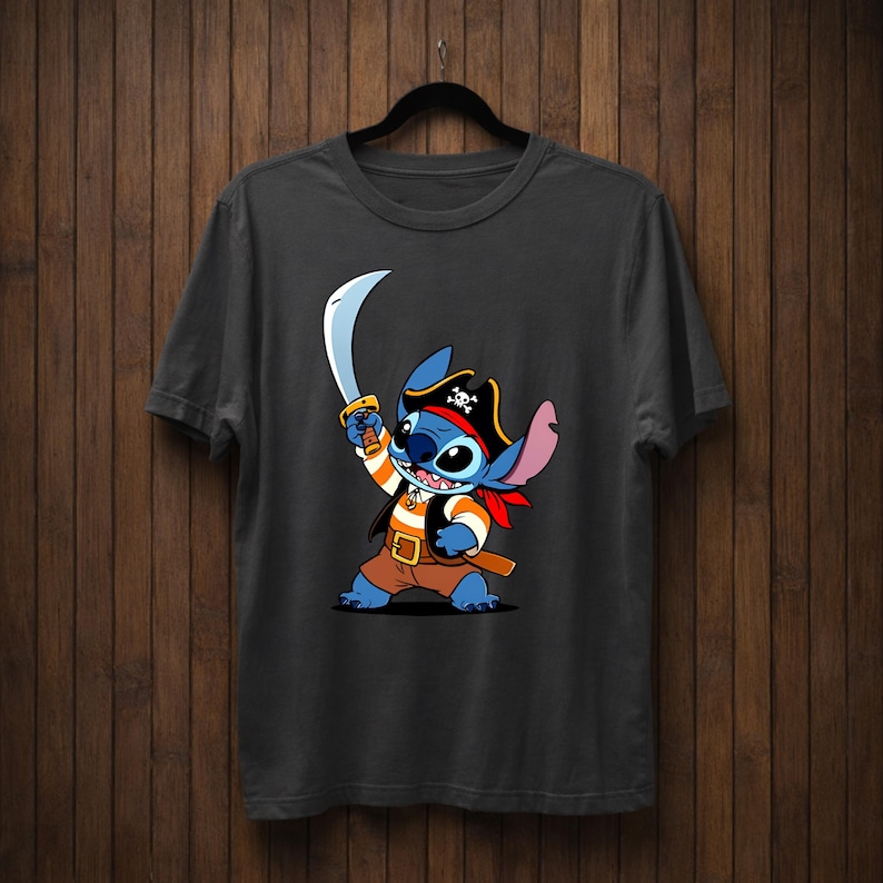 May include: Dark gray t-shirt featuring a cartoon Stitch character dressed as a pirate. Stitch is blue with large ears, wearing a pirate hat, holding a sword, and a red bandana. The shirt is hanging on a black hanger against a wooden background.