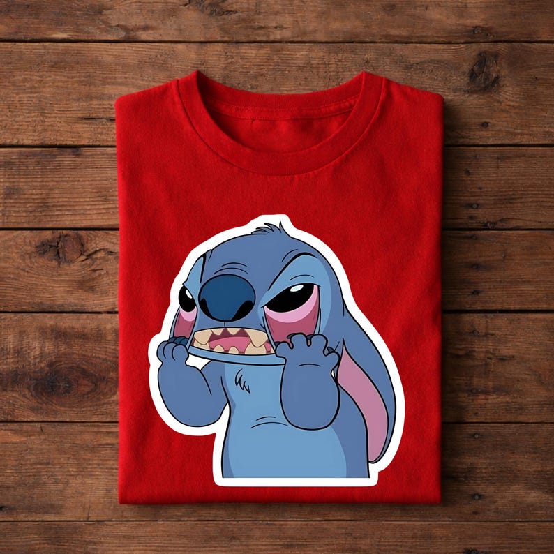May include: A red t-shirt featuring a cartoon image of Stitch from Lilo & Stitch. The image shows Stitch with an angry expression, with blue fur, large ears, and pink accents. The shirt is folded on a wooden surface.