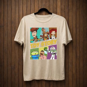 K&ouml;nnte beinhalten: Beiges T-Shirt mit einer farbenfrohen Grafik von Charakteren aus dem Animationsfilm "Toy Story". Das Design zeigt Woody, Buzz Lightyear und andere Charaktere in einem Raster, mit den Worten "TOY STORY" in Gelb.