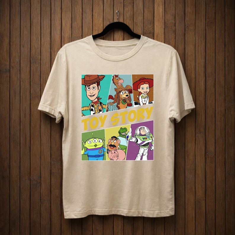 K&ouml;nnte beinhalten: Beiges T-Shirt mit einer farbenfrohen Grafik von Charakteren aus dem Animationsfilm "Toy Story". Das Design zeigt Woody, Buzz Lightyear und andere Charaktere in einem Raster, mit den Worten "TOY STORY" in Gelb.