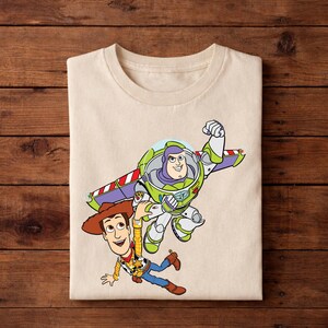 May include: A beige t-shirt featuring a graphic of Buzz Lightyear and Woody from Toy Story. Buzz Lightyear is in his spacesuit with wings extended, and Woody is in his cowboy outfit. The design is colorful and cartoon-like.
