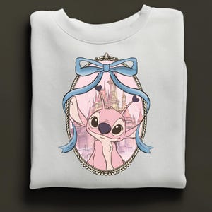 Puede incluir: Sudadera blanca con un dise&ntilde;o de personaje de dibujos animados rosa, enmarcado por una cinta azul y un castillo de fondo. El marco ovalado tiene un borde decorativo. El dise&ntilde;o est&aacute; centrado.