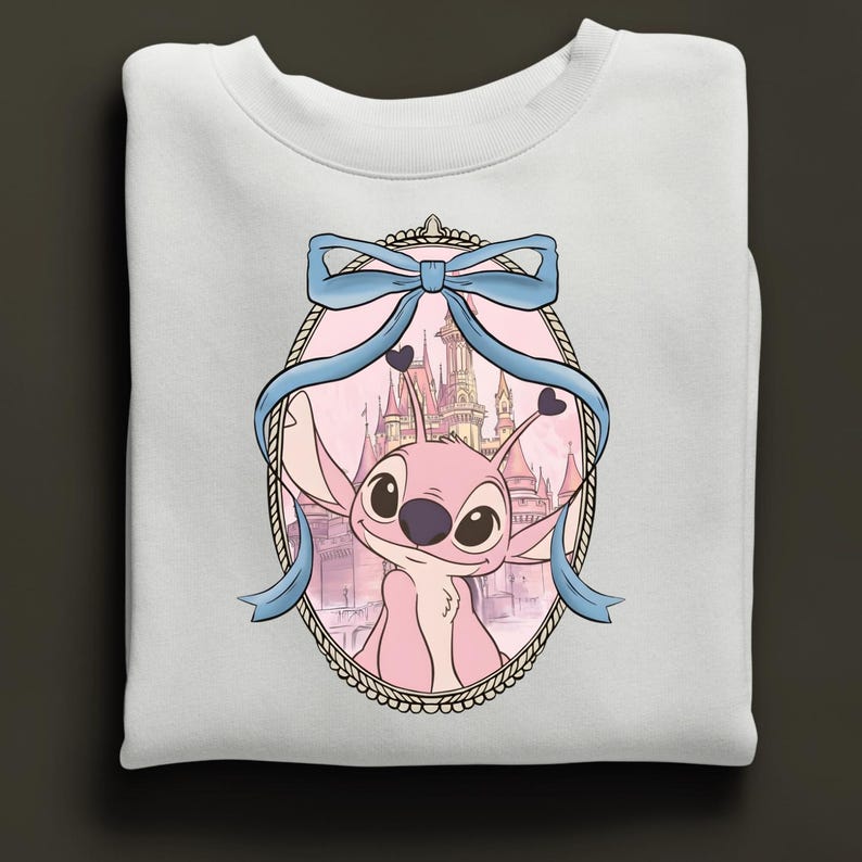 Puede incluir: Sudadera blanca con un dise&ntilde;o de personaje de dibujos animados rosa, enmarcado por una cinta azul y un castillo de fondo. El marco ovalado tiene un borde decorativo. El dise&ntilde;o est&aacute; centrado.